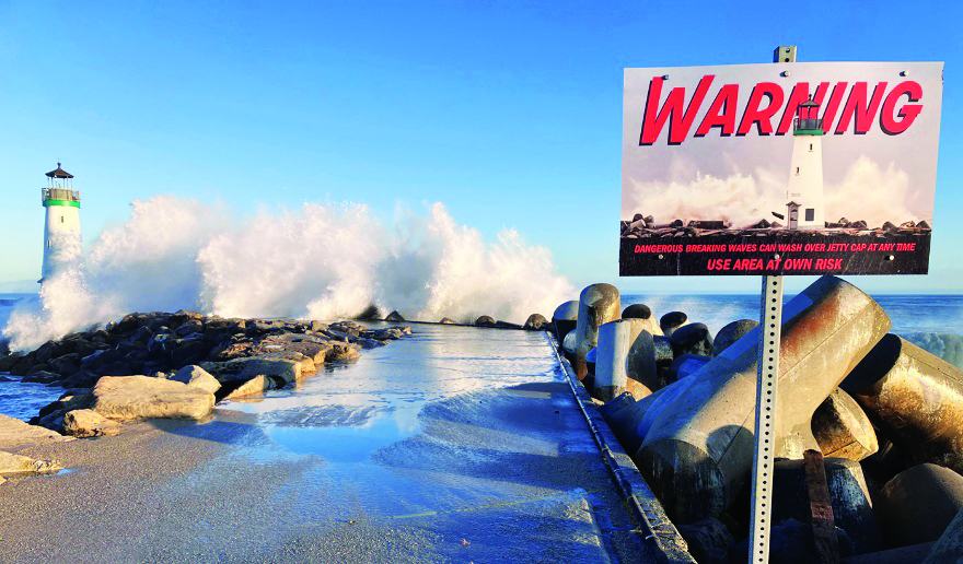Massive wave crashes over the Santa Cruz Harbor jetty beside a warning sign and lighthouse.