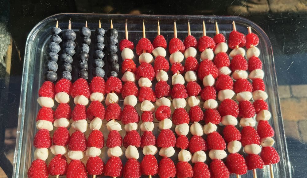 Fruit skewers arranged like an American flag with blueberries, raspberries and white cheese or melon in a glass dish.