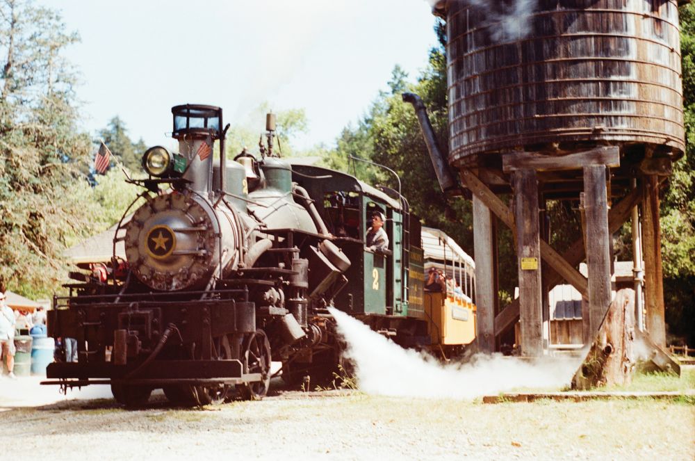 Historic steam locomotive Engine No. 2 at Roaring Camp Railroad releases steam beside a wooden water tower.