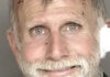 Violent Felon Runs Police booking photo of an older bearded man with white gauze near his right ear.