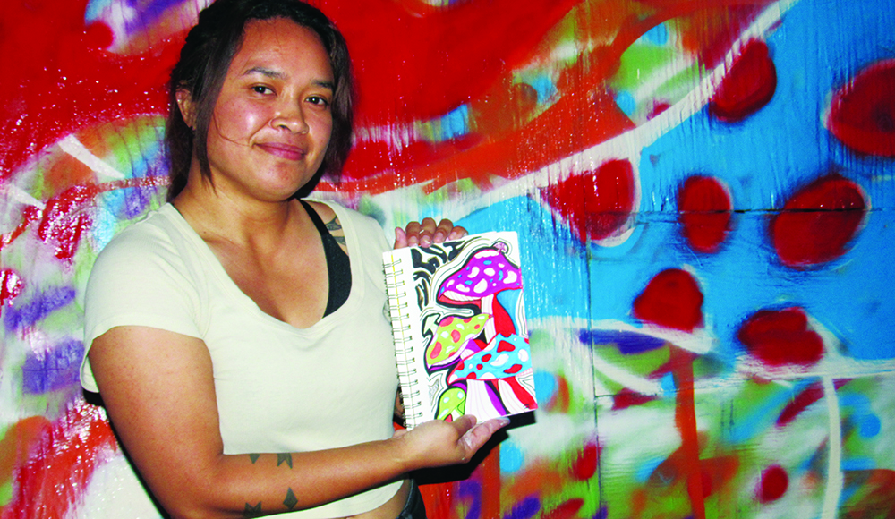 Artist Joyleen Torwan holds a colorful mushroom drawing in front of a painted mural.
