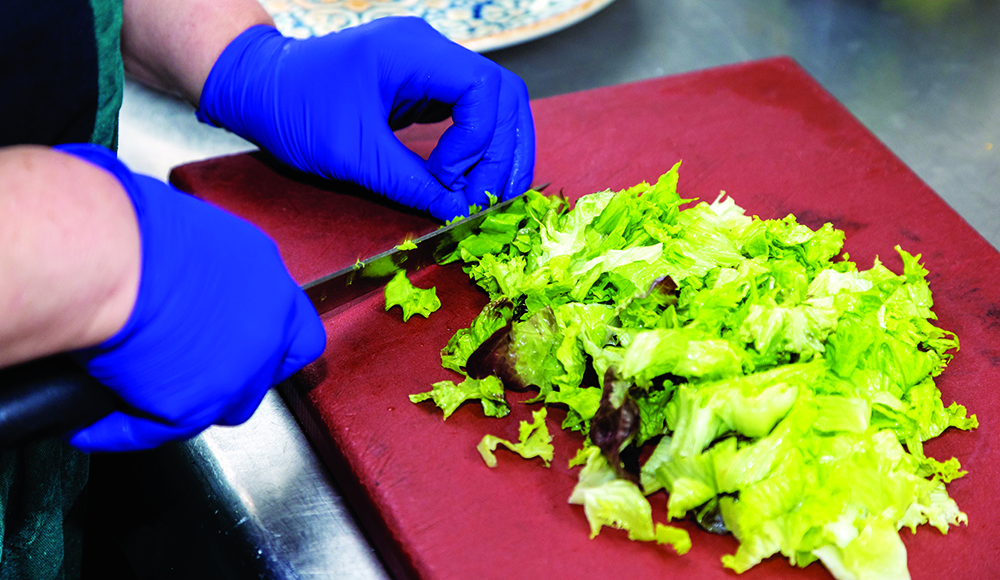 A food worker wearing blue gloves chops fresh lettuce on a cutting board.