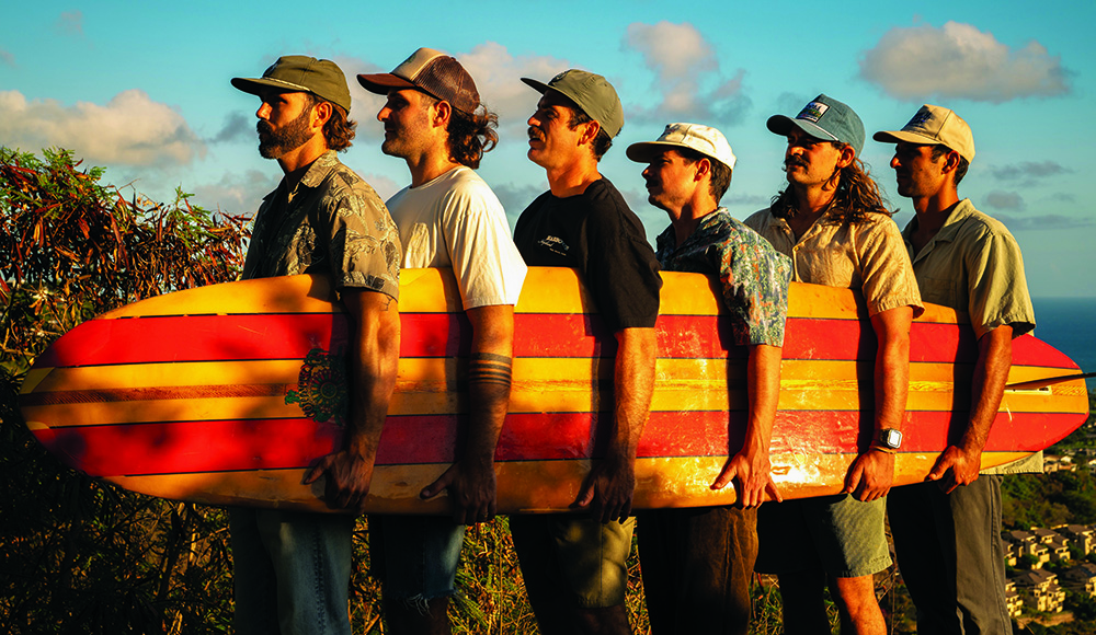 Twin Fin band members carrying a vintage surfboard outdoors.