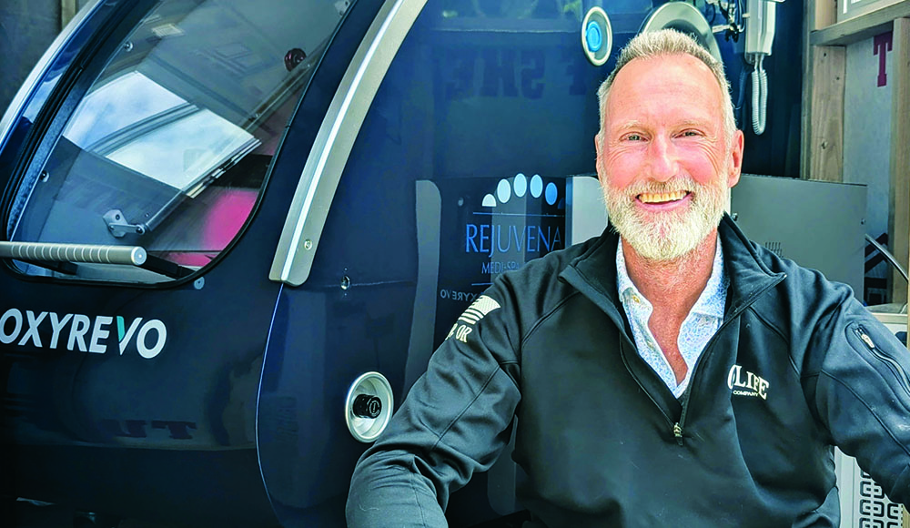 Dr. Jay Pennock sits beside a hyperbaric oxygen therapy chamber at Rejuvenate Medi-Spa and Wellness Center.
