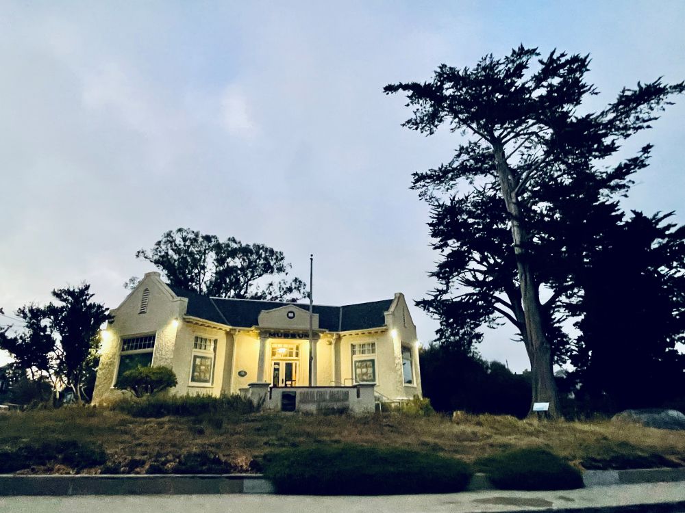 Museum building illuminated at dusk beneath a large cypress tree.