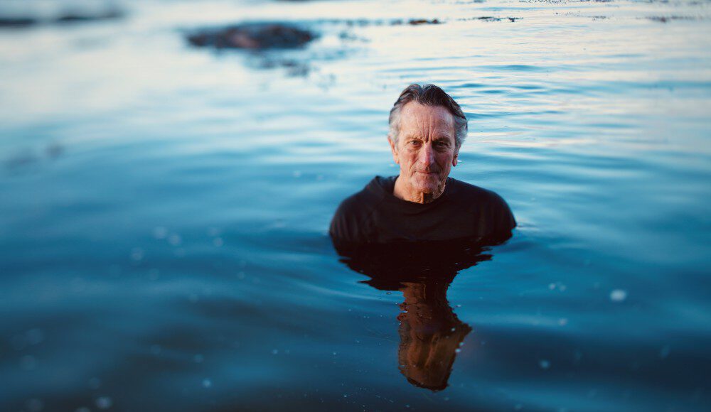 Older surfer standing chest-deep in calm blue ocean water, wearing a black shirt.