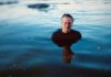 Neoprene Noir Older surfer standing chest-deep in calm blue ocean water, wearing a black shirt.