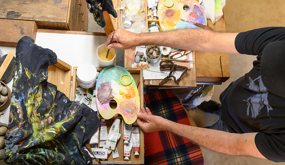 Ea Eckerman holds a colorful painter’s palette and dips a brush into a cup at a workbench crowded with art supplies.