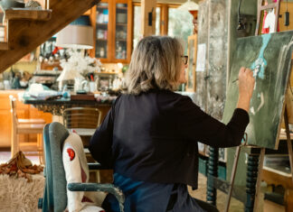 A Three-Week Vision Quest Dee Hooker sits at an easel, painting a figure on a dark canvas in a home studio beneath a wooden staircase.