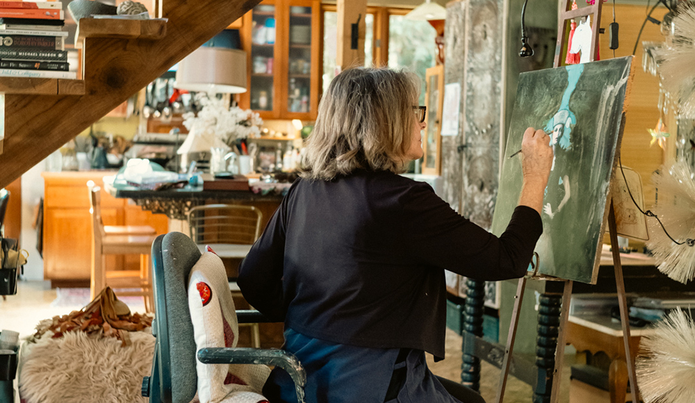 Dee Hooker sits at an easel, painting a figure on a dark canvas in a home studio beneath a wooden staircase.