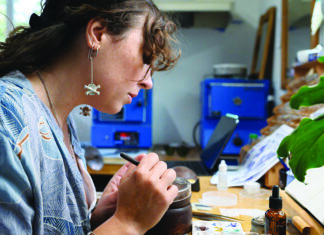 Darci Shea Bogdan works with a fine brush at a studio bench beside sketches, small containers and a leafy plant.