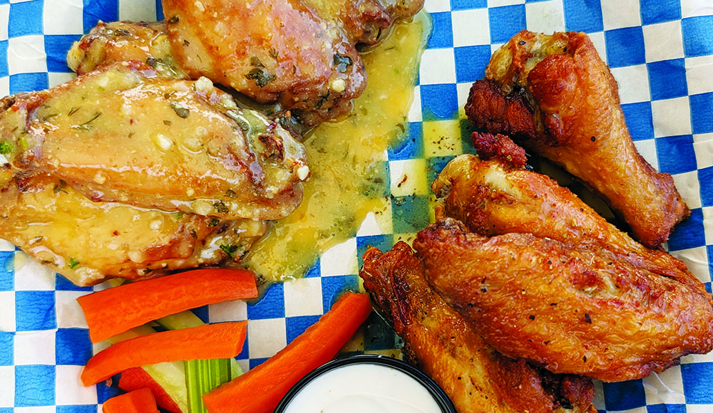 Chicken wings from Evil Wings served on blue-and-white checkered paper with carrot sticks, celery and a cup of dipping sauce.