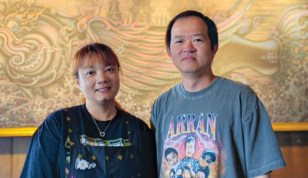 Nancy and Arun Sadkiriyasab stand together inside Lemongrass restaurant in Moss Landing, with gold artwork behind them.