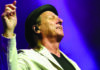 Crimson at The Quarry Adrian Belew raises one hand onstage, wearing a black hat and light-colored blazer against purple stage lighting.