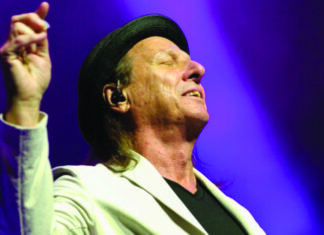 Crimson at The Quarry Adrian Belew raises one hand onstage, wearing a black hat and light-colored blazer against purple stage lighting.