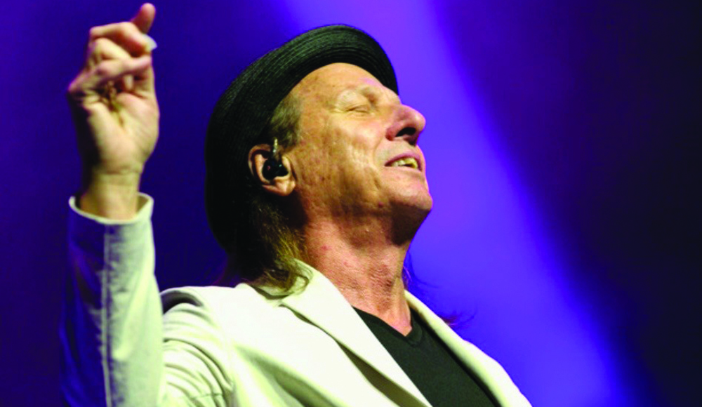 Adrian Belew raises one hand onstage, wearing a black hat and light-colored blazer against purple stage lighting.