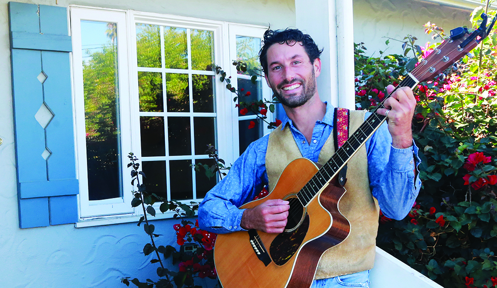 Alt text: Santa Cruz Porchfest organizer Darrow Feldstein plays an acoustic guitar on a porch beside flowering roses.