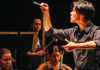 Distinguished Artists Carlin Truong conducts with a raised baton and an outstretched hand as string musicians perform behind him.