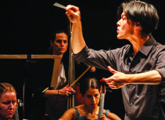 Distinguished Artists Carlin Truong conducts with a raised baton and an outstretched hand as string musicians perform behind him.