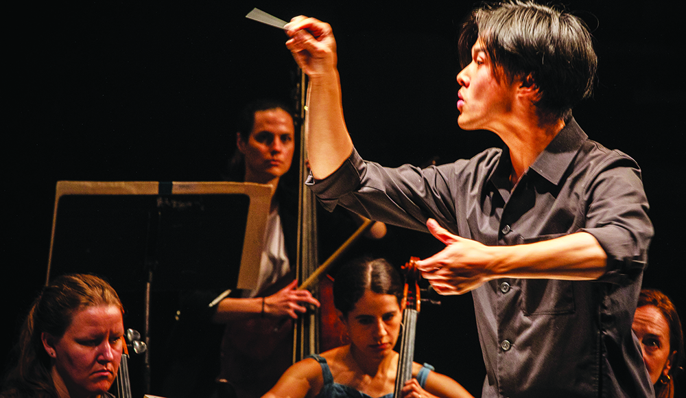 Carlin Truong conducts with a raised baton and an outstretched hand as string musicians perform behind him.