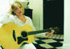 Things to do in Santa Cruz Liz Longley in a white shirt, seated with an acoustic guitar beside a black-and-white tiled floor.