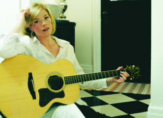 Things to do in Santa Cruz Liz Longley in a white shirt, seated with an acoustic guitar beside a black-and-white tiled floor.
