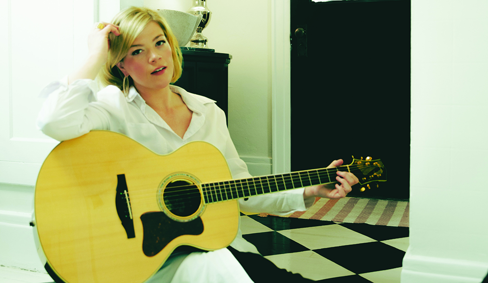 Liz Longley in a white shirt, seated with an acoustic guitar beside a black-and-white tiled floor.