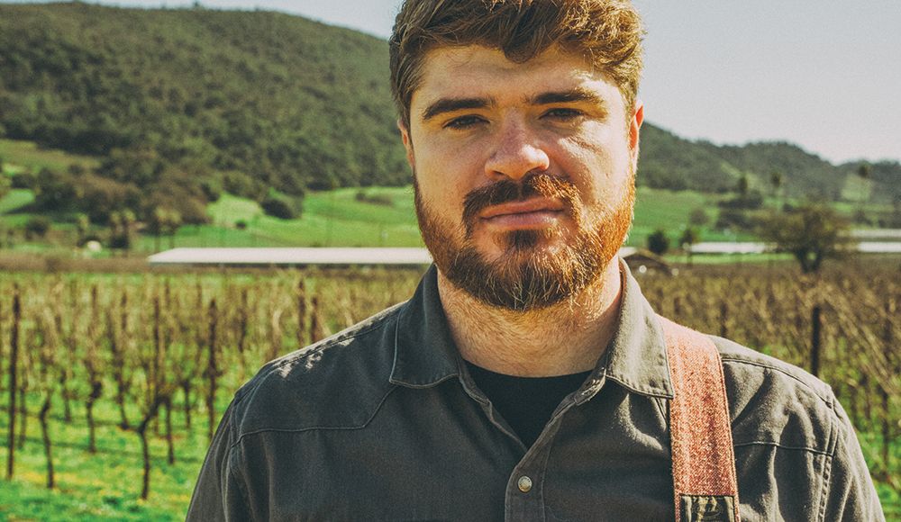 Marty O’Reilly wearing a guitar strap, with vineyard rows and green hills behind him.