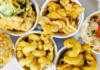 Dairy Departed Overhead view of six sample cups of vegan mac and cheese, with varied pasta shapes and toppings including broccoli and peppers.