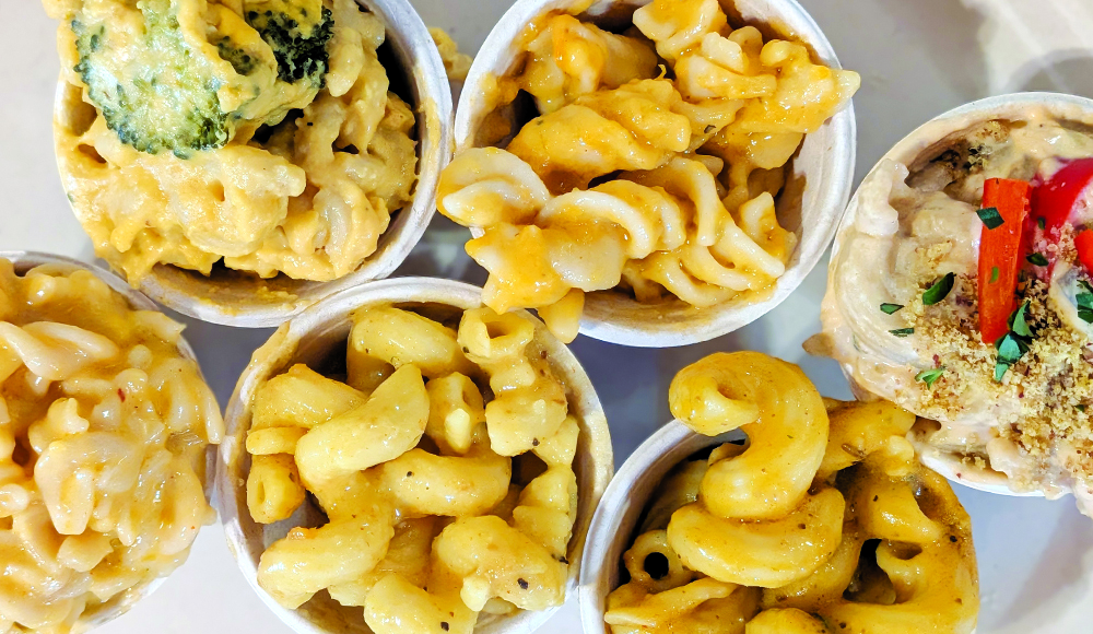 Overhead view of six sample cups of vegan mac and cheese, with varied pasta shapes and toppings including broccoli and peppers.