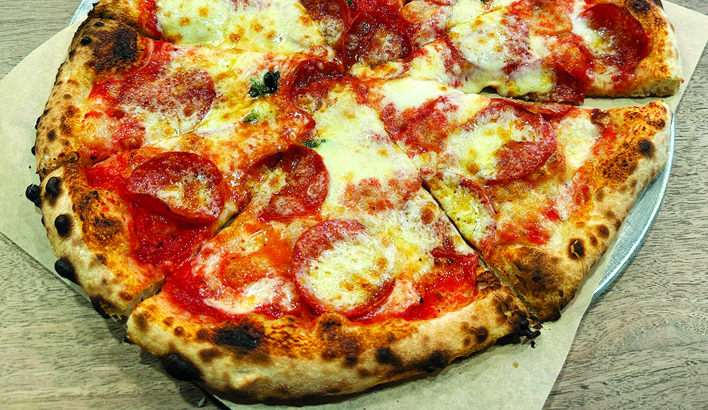 Sliced pizza topped with tomato sauce, melted cheese and rounds of salami, pepperoni, and peppers with a blistered crust on a metal tray.