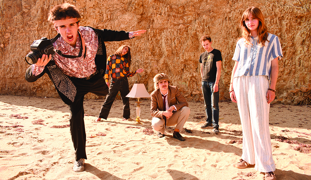 Five Coyote Theater band members pose on sand in front of a rocky bluff, with a camera and floor lamp as props.