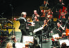Season of Sound A conductor holds a baton and extends one arm toward orchestral musicians playing strings on a concert stage.
