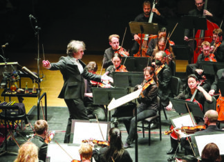 Season of Sound A conductor holds a baton and extends one arm toward orchestral musicians playing strings on a concert stage.