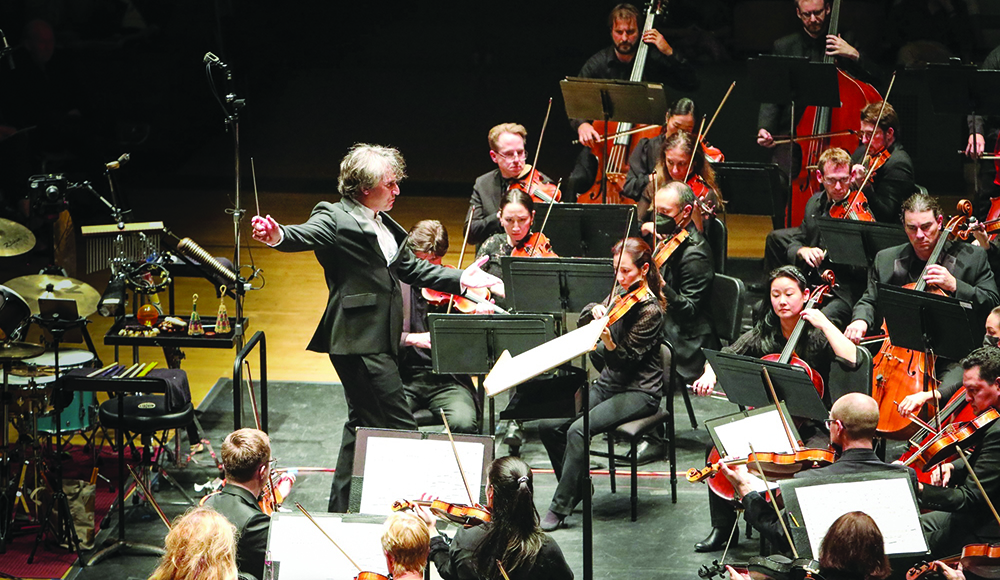 A conductor holds a baton and extends one arm toward orchestral musicians playing strings on a concert stage.