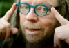 Reality, Remade Jaron Lanier points toward his round blue glasses in a close-up outdoor portrait.