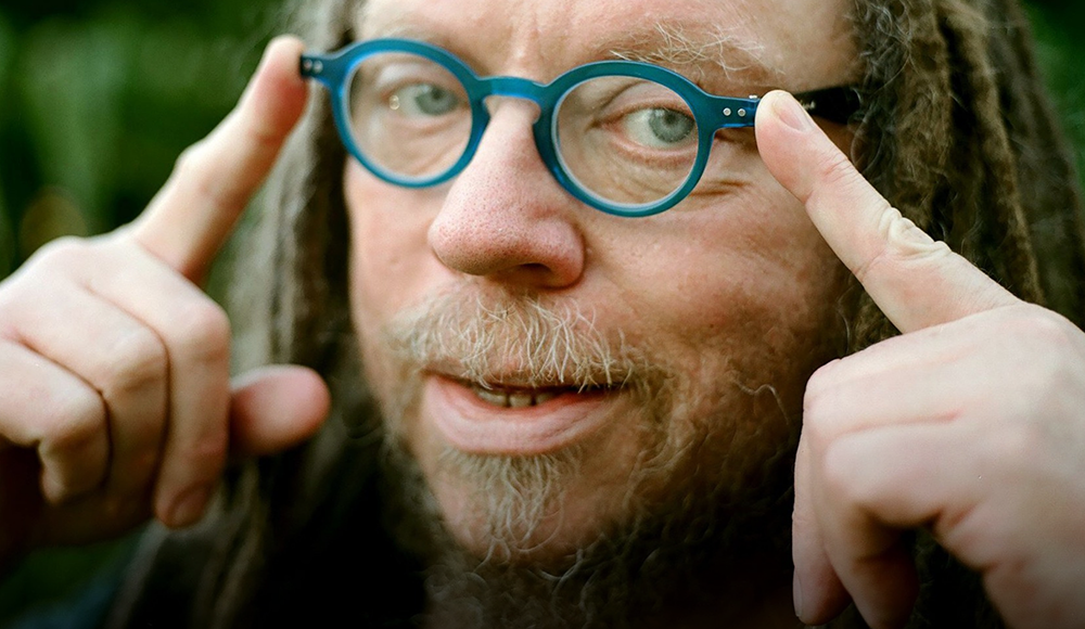 Jaron Lanier points toward his round blue glasses in a close-up outdoor portrait.