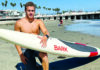 There and Back Again Oliver Levine kneels on a beach holding a white Bark paddleboard, with a pier and palm trees behind him.
