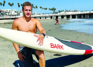 There and Back Again Oliver Levine kneels on a beach holding a white Bark paddleboard, with a pier and palm trees behind him.