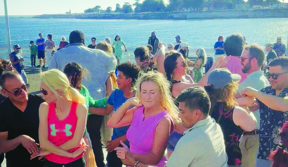 Couples dance close together on Santa Cruz Wharf, with the sunlit ocean and West Cliff coastline behind them.
