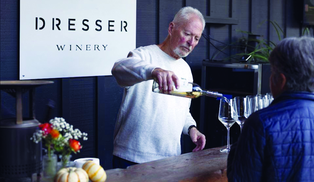 A man pours white wine into a tasting glass for a guest at a counter beneath a Dresser Winery sign.