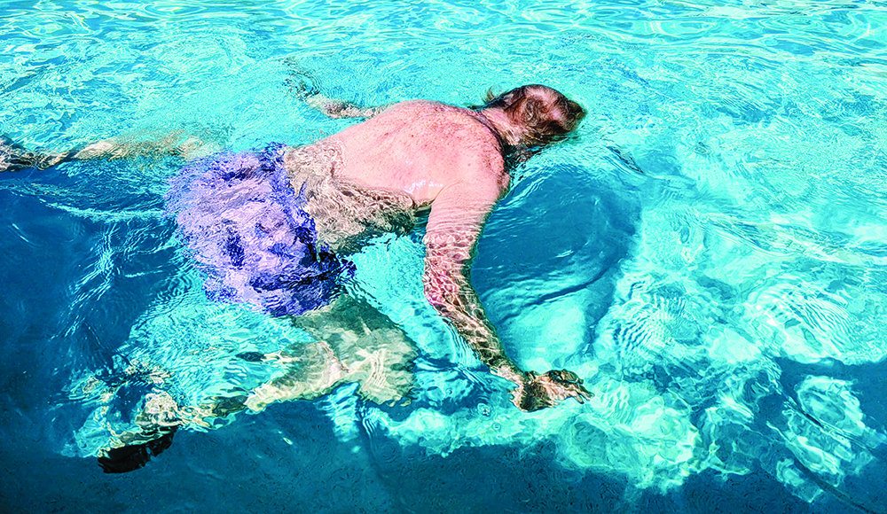 Ed Frey floats face-down with his arms extended in the sunlit pool at Simpkins Family Swim Center.