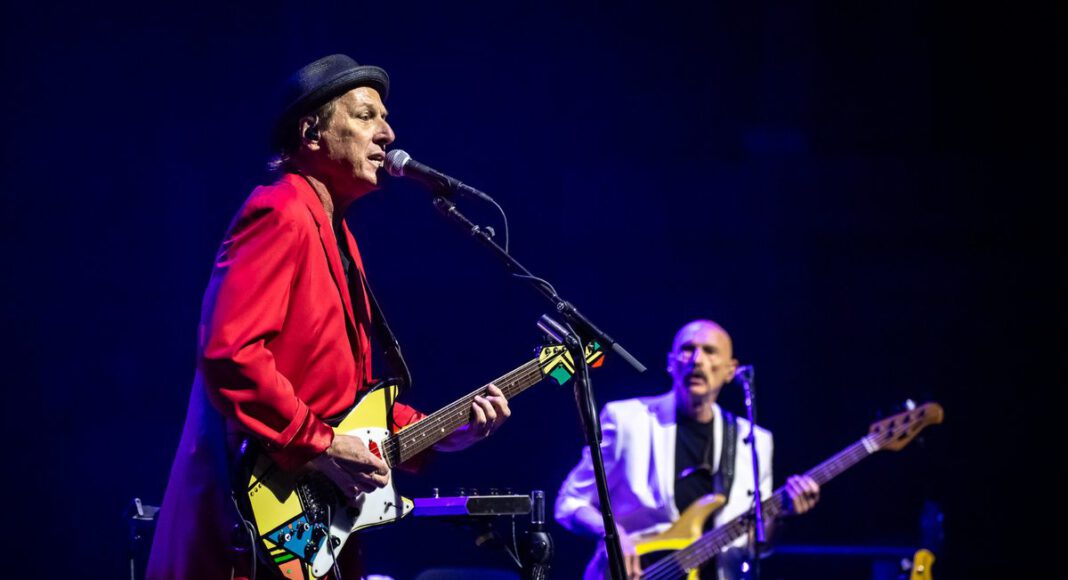 Adrian Belew sings and plays guitar in a red suit, with bassist Tony Levin behind him onstage.