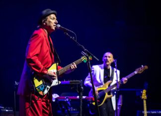 Crimson Roars in the Redwoods Adrian Belew sings and plays guitar in a red suit, with bassist Tony Levin behind him onstage.