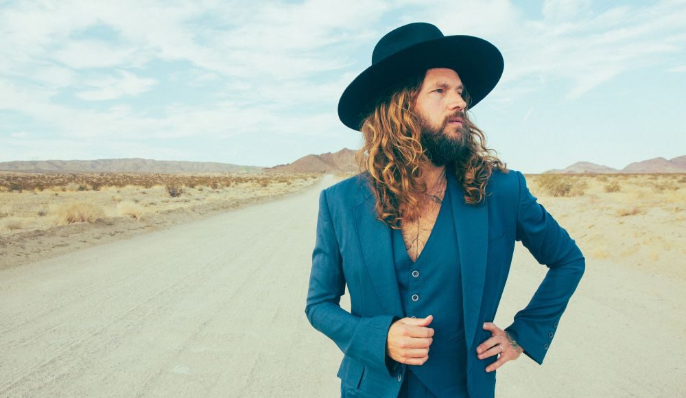 Jay Buchanan wears a blue suit and wide-brimmed black hat on a desert road, with mountains in the distance.