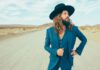 Dazzling from the Desert Jay Buchanan wears a blue suit and wide-brimmed black hat on a desert road, with mountains in the distance.