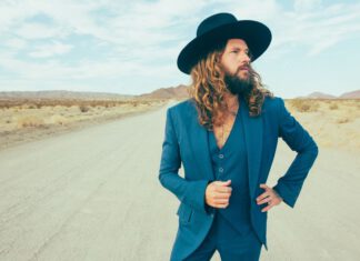 Dazzling from the Desert Jay Buchanan wears a blue suit and wide-brimmed black hat on a desert road, with mountains in the distance.