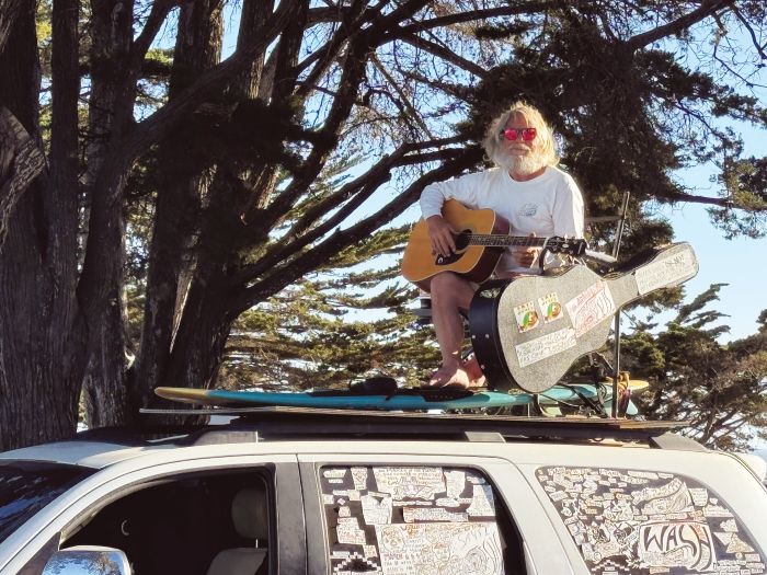 Barefoot guitarist wearing red sunglasses sits atop a car beside a sticker-covered guitar case beneath spreading trees.